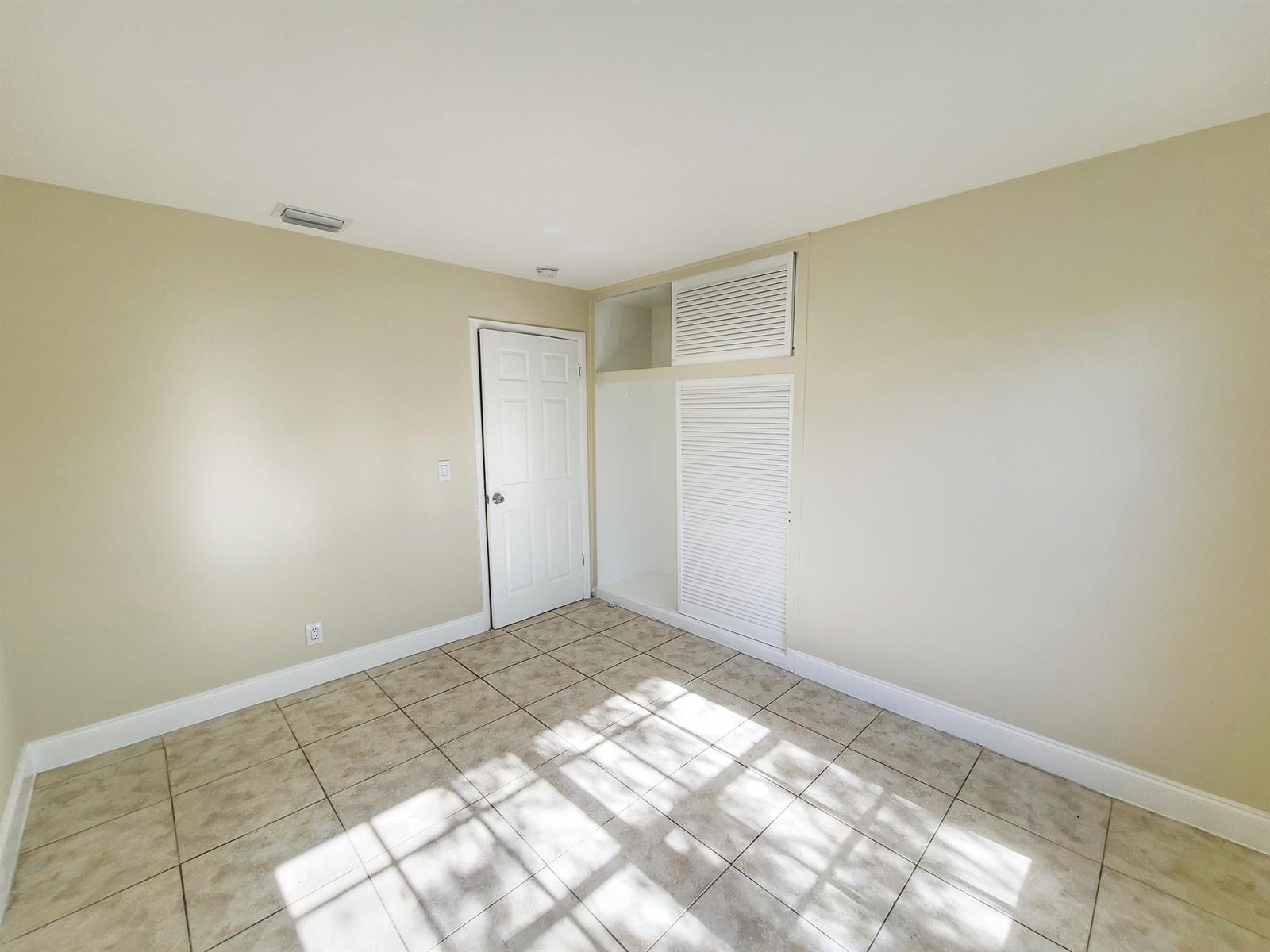 An empty room features beige walls, tiled flooring, a white door, and built-in louvered closets — 420 Blue Heron Boulevard # 2, West Palm Beach FL 33404, WEST PALM BEACH, FL