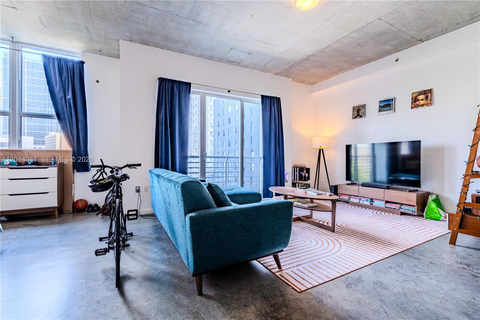 A living room features a blue sofa, a television, a bicycle, and concrete ceilings — 133 NE 2nd Ave # 609, Miami FL 33132, MIAMI, FL