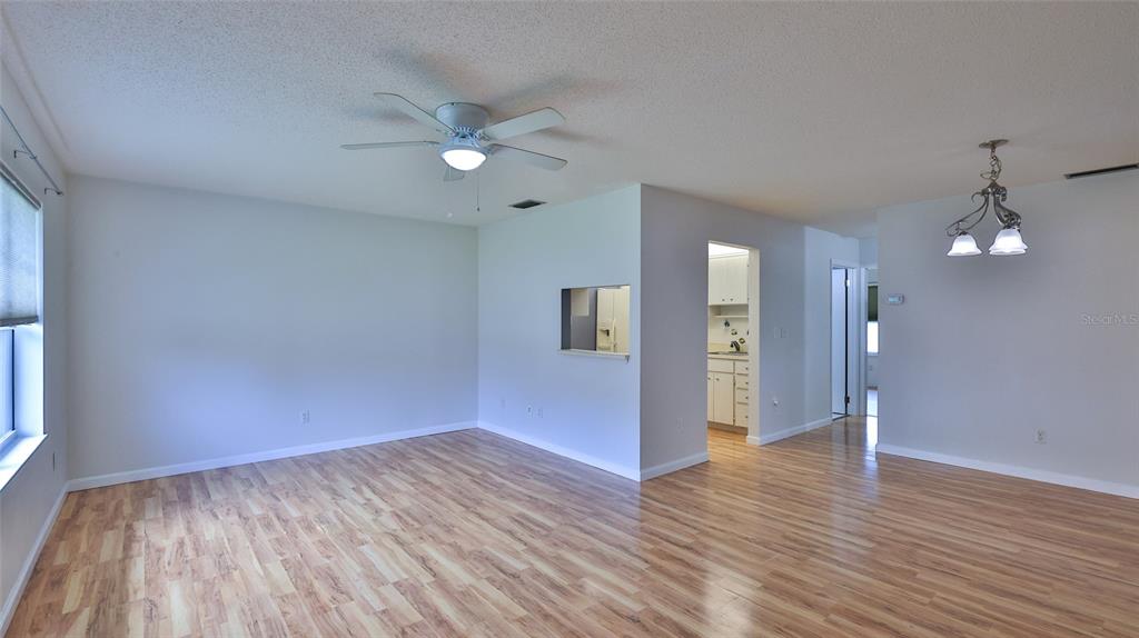 An empty living room features light wood-look flooring, a ceiling fan, and a pass-through window — 12 BOGEY CIRCLE, NEW SMYRNA BEACH, FL