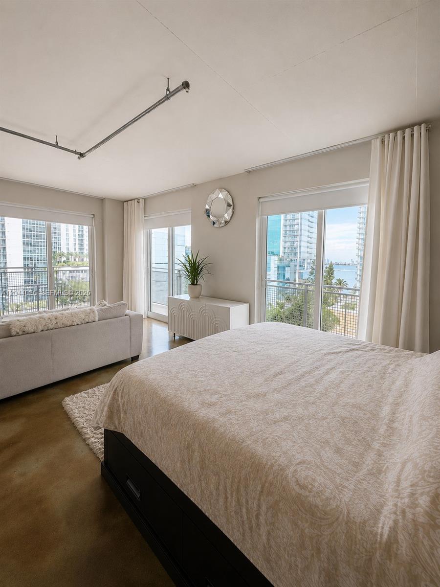 A bedroom features a bed, sofa, concrete floors, and large windows overlooking a city skyline — 455 NE 25th St # 806, Miami FL 33137, MIAMI, FL