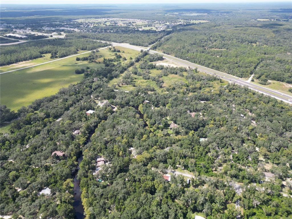 An aerial view shows a wooded residential area with a winding creek and a nearby highway — 0012 MARCHMONT CIRCLE, DADE CITY, FL