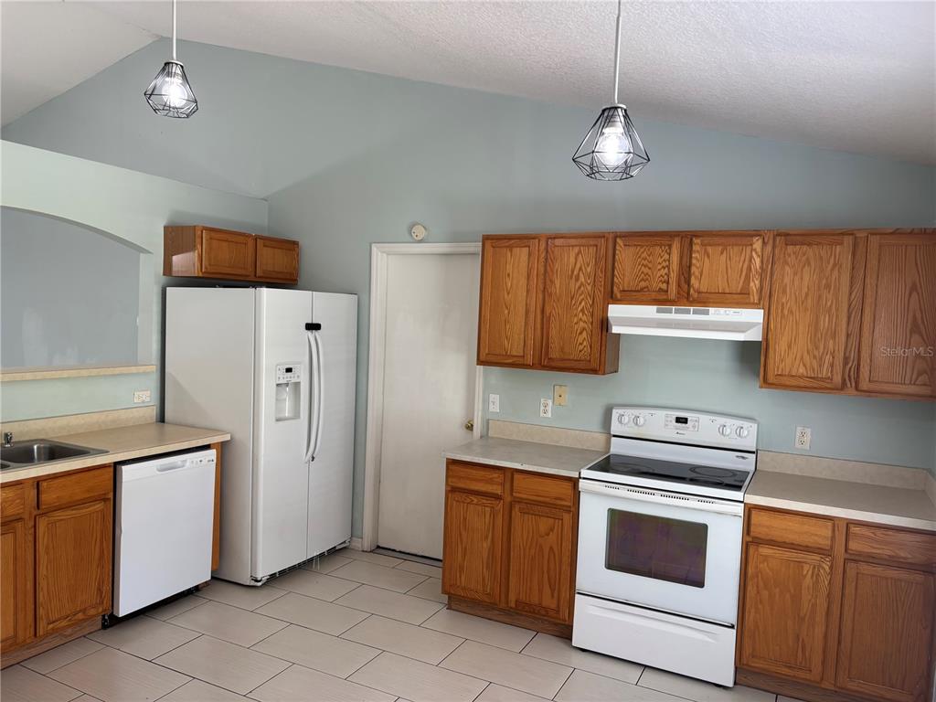 A kitchen features light blue walls, oak cabinets, white appliances, and two geometric pendant lights — 120 SPOONBILL COURT, POINCIANA, FL