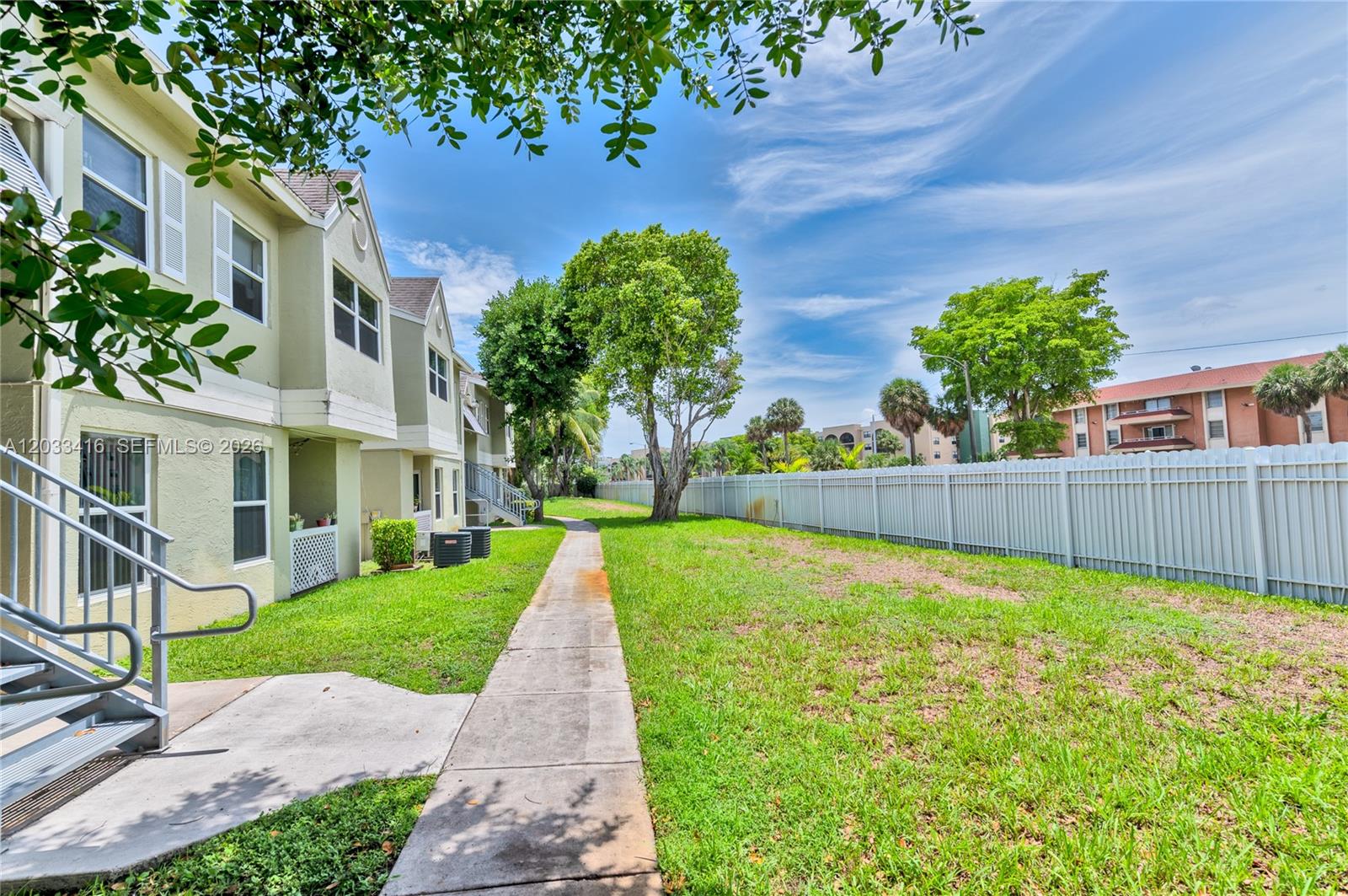 A sidewalk runs alongside a multi-story apartment building with a white fence and lawn — 17485 NW 67th Ct # 21, Hialeah FL 33015, HIALEAH, FL