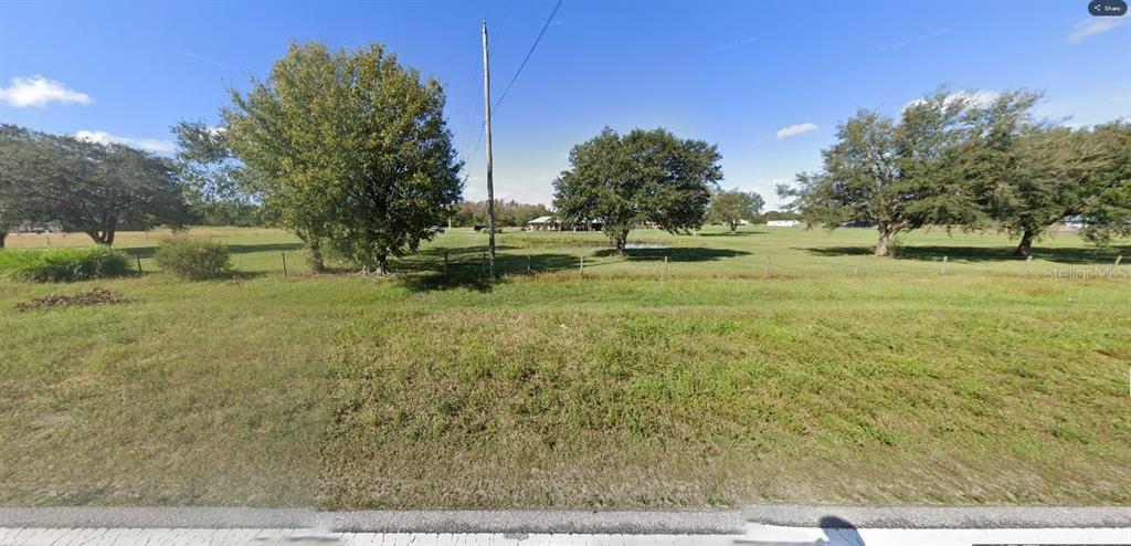 A grassy, fenced lot features several large trees and a distant house under a blue sky — OLD GRADE RD, POLK CITY, FL