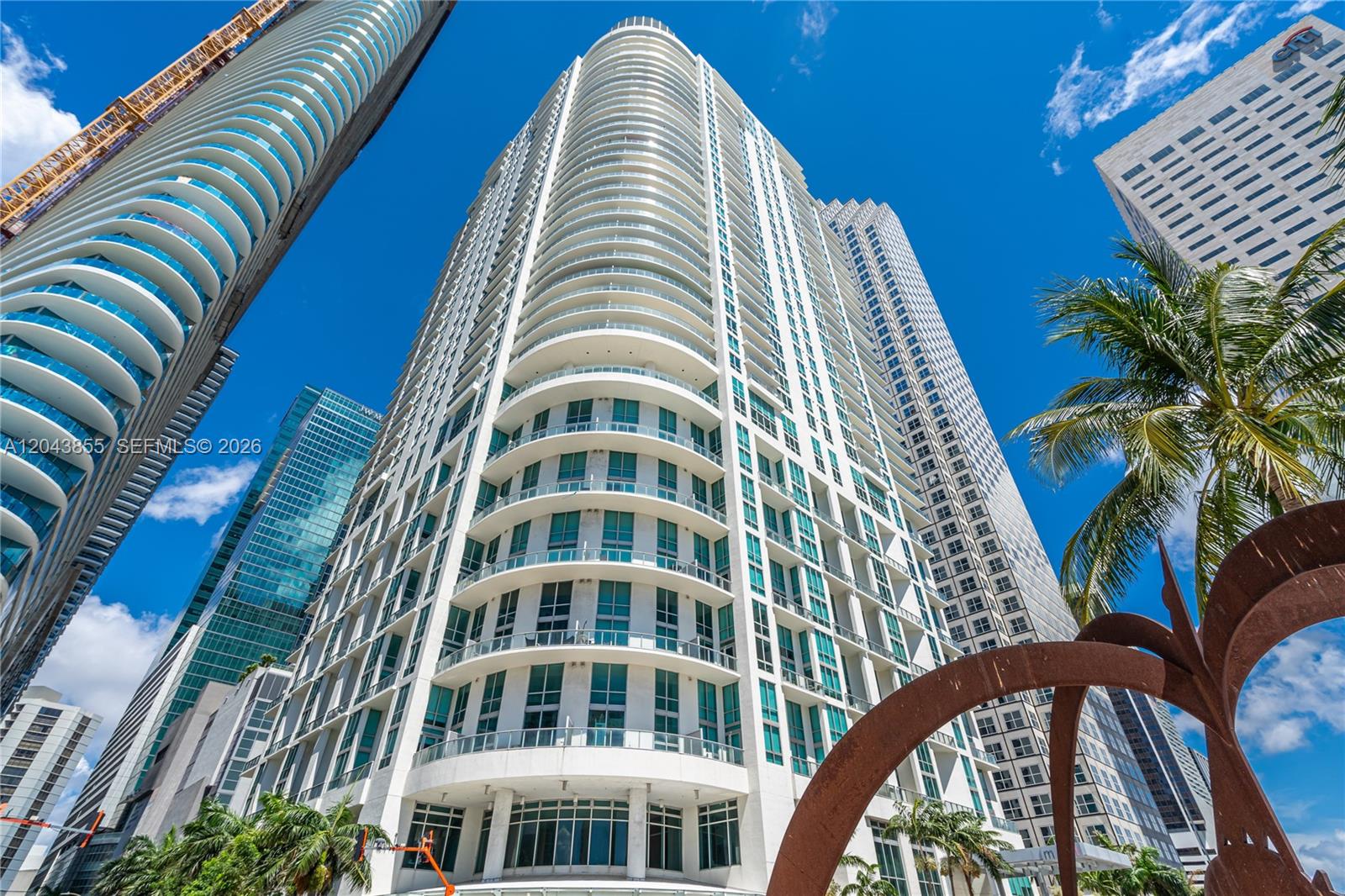 A balcony overlooks a waterfront walkway, palm trees, and high-rise buildings under a blue sky — 300 S Biscayne Blvd # 612, Miami FL 33131, MIAMI, FL