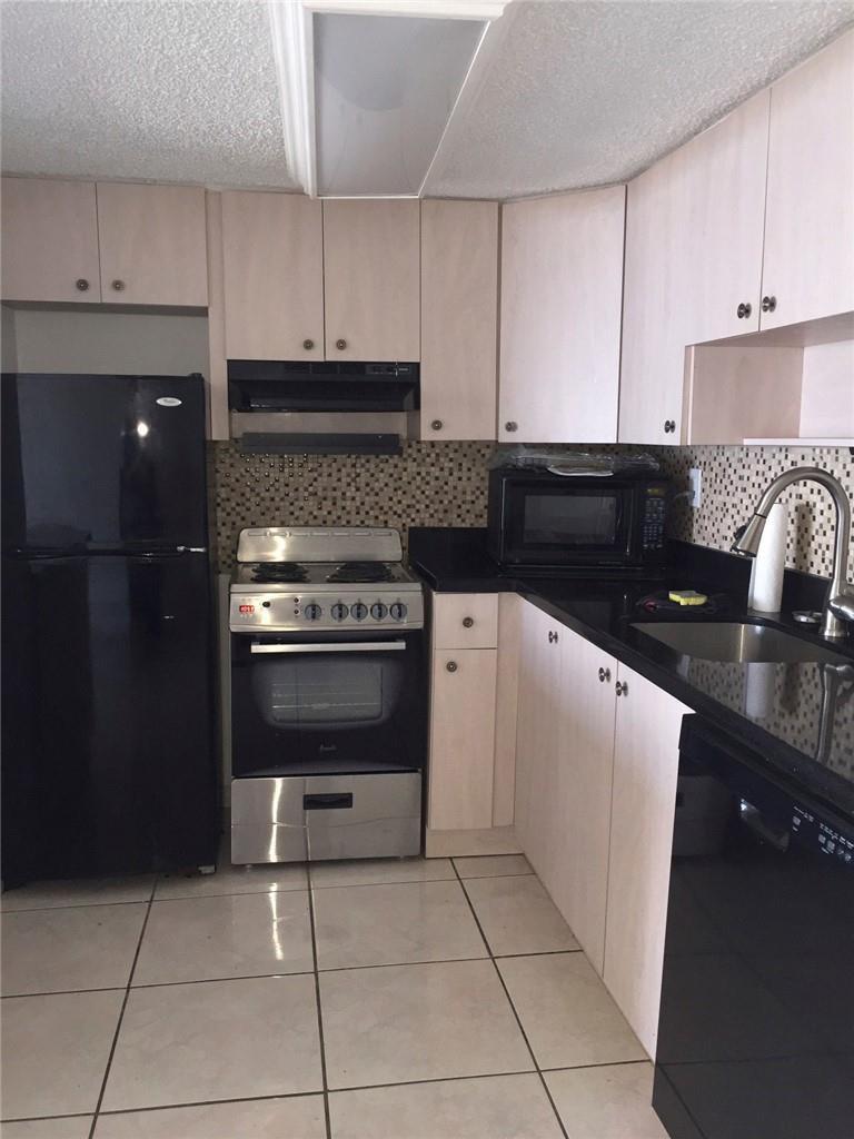A kitchen features light wood cabinets, black countertops, a black refrigerator, and a tiled floor — 300 E Oregon Street E # 303, Hollywood FL 33019, HOLLYWOOD, FL