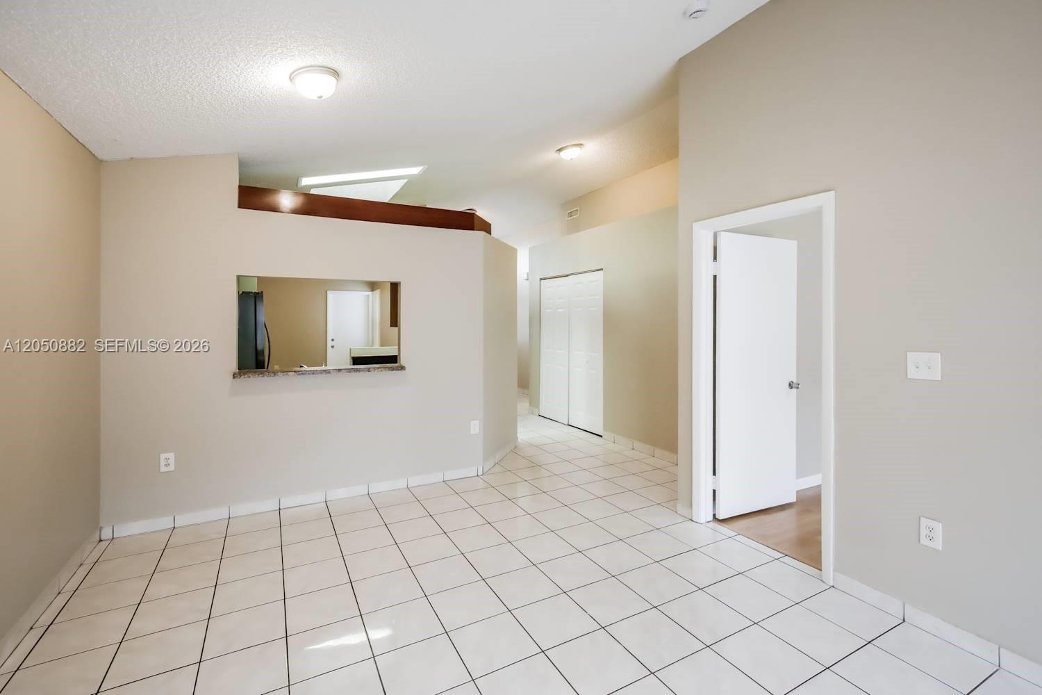 An empty room features white tile flooring, beige walls, and a pass-through window to the kitchen — CUTLER BAY, FL