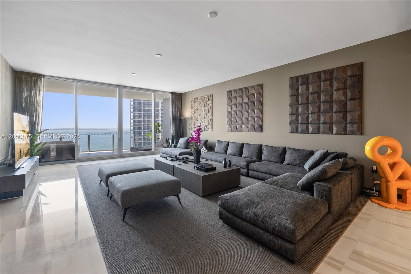 A modern living room features a large gray sectional sofa, matching ottomans, and floor-to-ceiling windows — 300 Biscayne Blvd Way # 3104, Miami FL 33131, MIAMI, FL