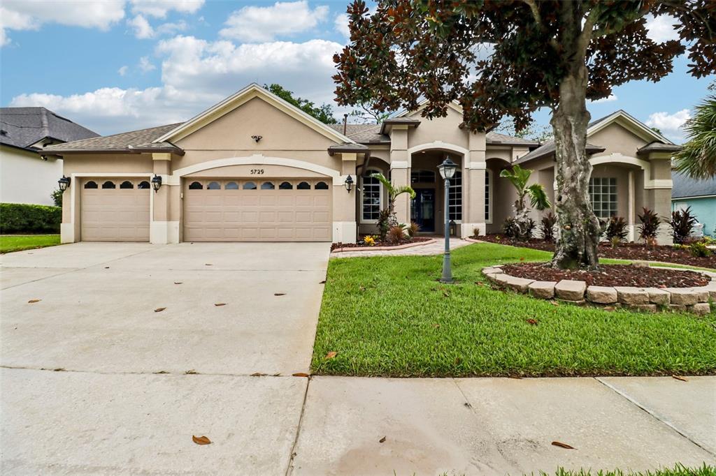 A single-story suburban house with a three-car garage, tan stucco exterior, and a landscaped yard — 5729 OAK LAKE TRAIL, OVIEDO, FL