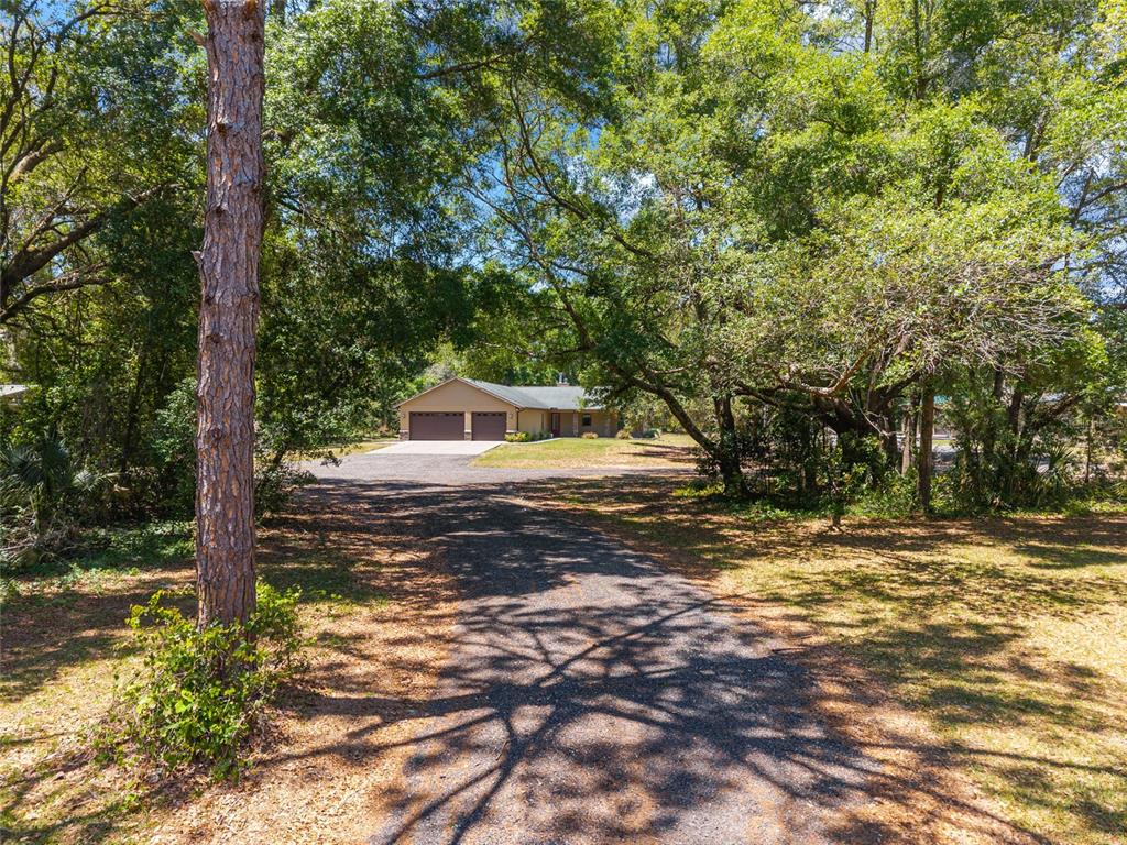 A gravel driveway leads to a single-story house with a two-car garage among mature trees — 5190 NW 90TH AVENUE, CHIEFLAND, FL