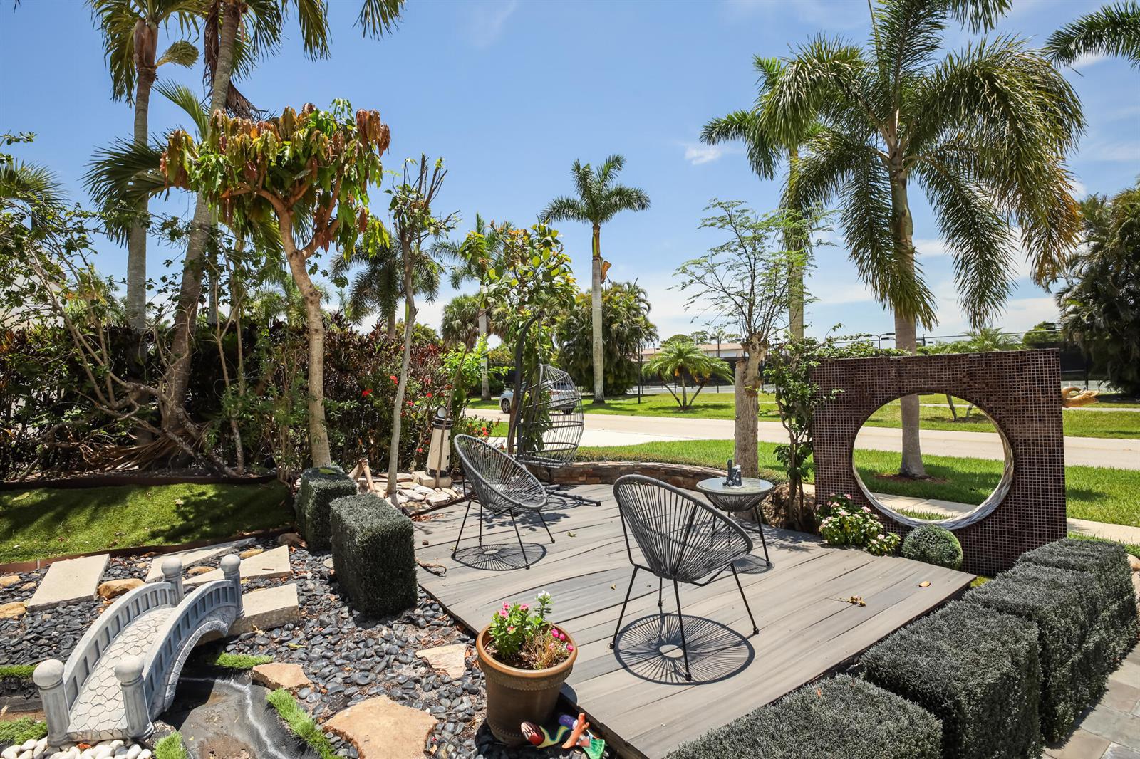 A backyard patio features a wooden deck, modern chairs, a small bridge, and palm trees — LAKE WORTH, FL