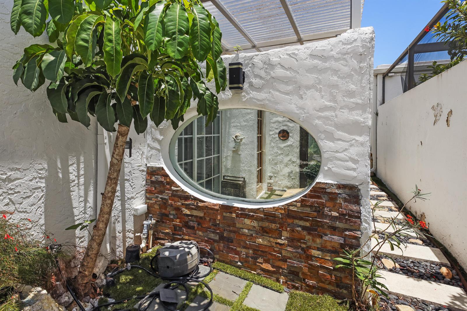 An outdoor patio features a textured white wall with a large oval mirror and stone base — LAKE WORTH, FL