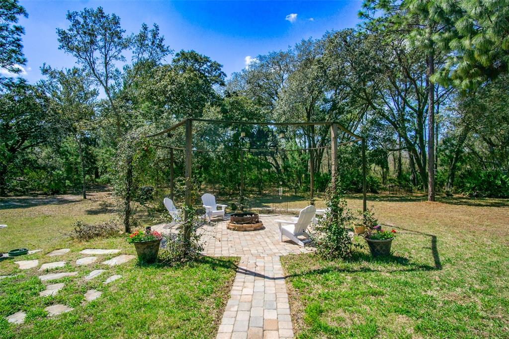 A stone patio with a fire pit and pergola sits in a grassy, wooded backyard — 11092 MIRAGE AVENUE, WEEKI WACHEE, FL