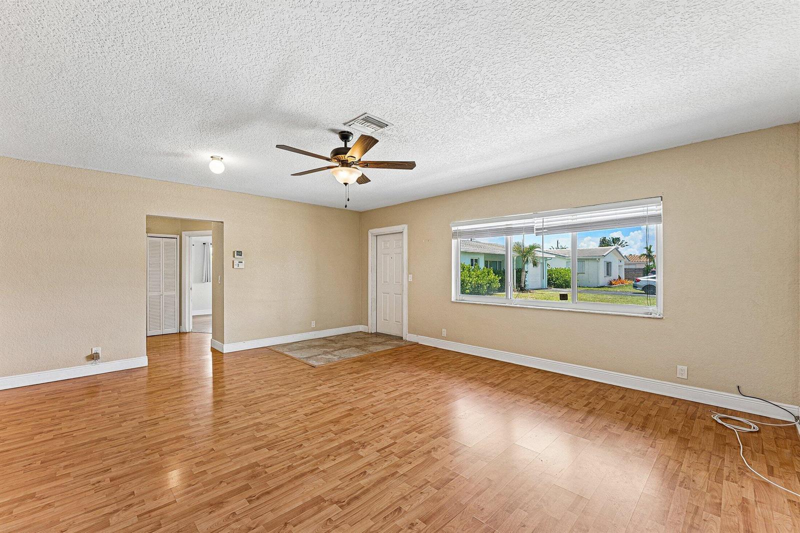 An empty living room features wood-look flooring, a ceiling fan, and a large front window — 301 SE 3rd Street, Deerfield Beach FL 33441, DEERFIELD BEACH, FL