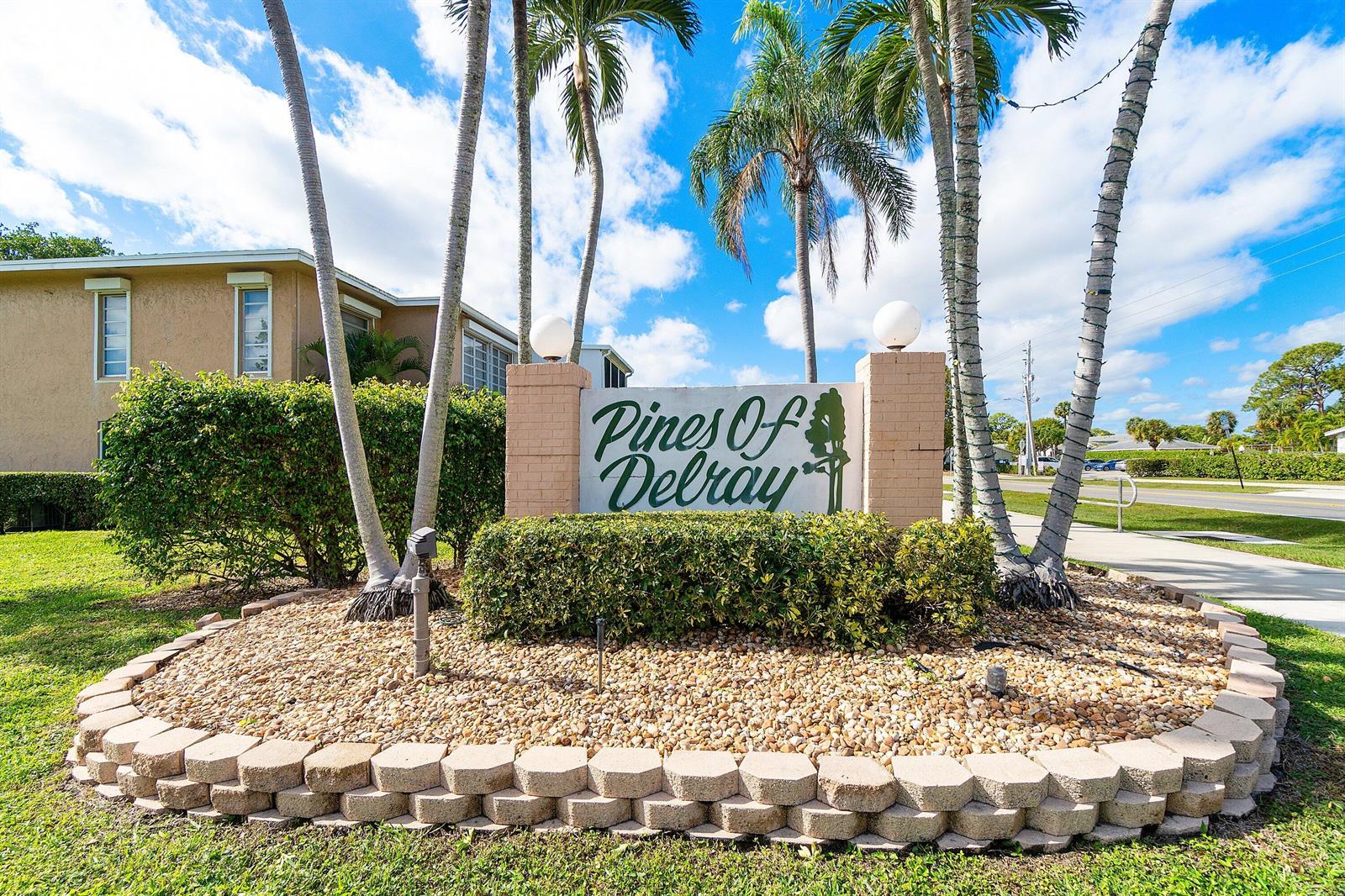 A stone sign for Pines of Delray stands in a landscaped bed with palm trees — 1020 Silk Oak Terrace # 102, Delray Beach FL 33445, DELRAY BEACH, FL