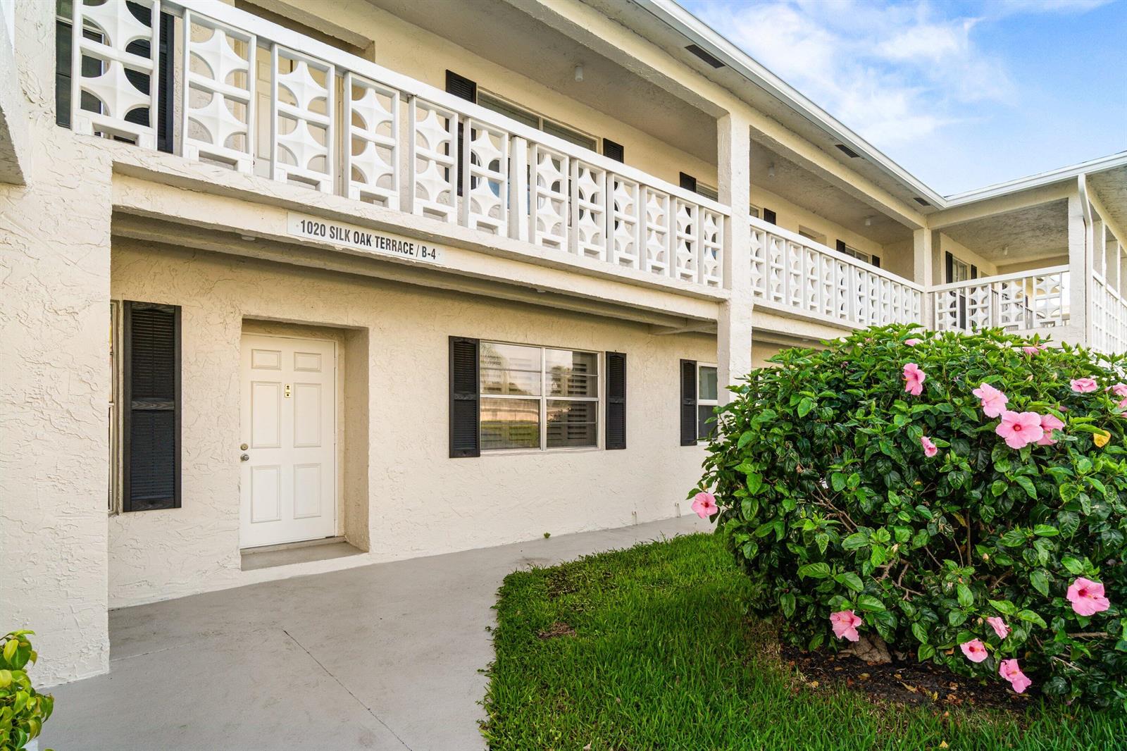A two-story apartment building exterior features a white stucco facade, decorative railings, and a hibiscus bush — 1020 Silk Oak Terrace # 102, Delray Beach FL 33445, DELRAY BEACH, FL