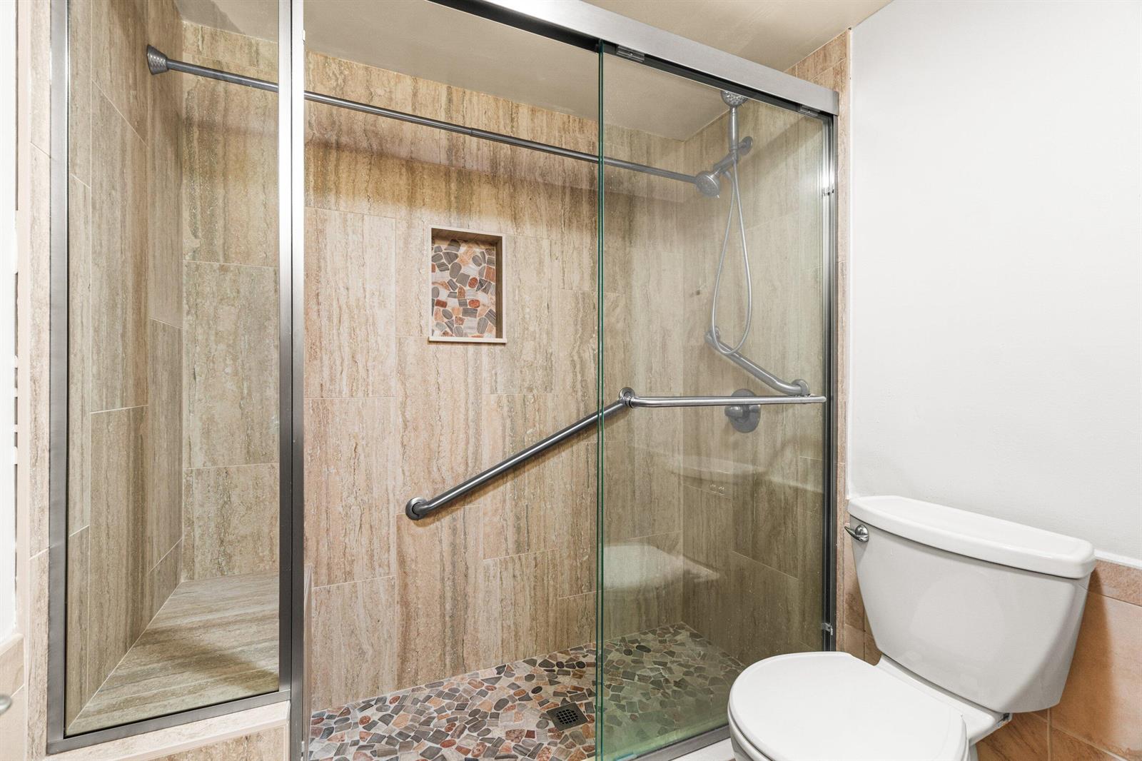 A bathroom features a walk-in shower with stone tile walls, a pebble floor, and grab bars — 1020 Silk Oak Terrace # 102, Delray Beach FL 33445, DELRAY BEACH, FL