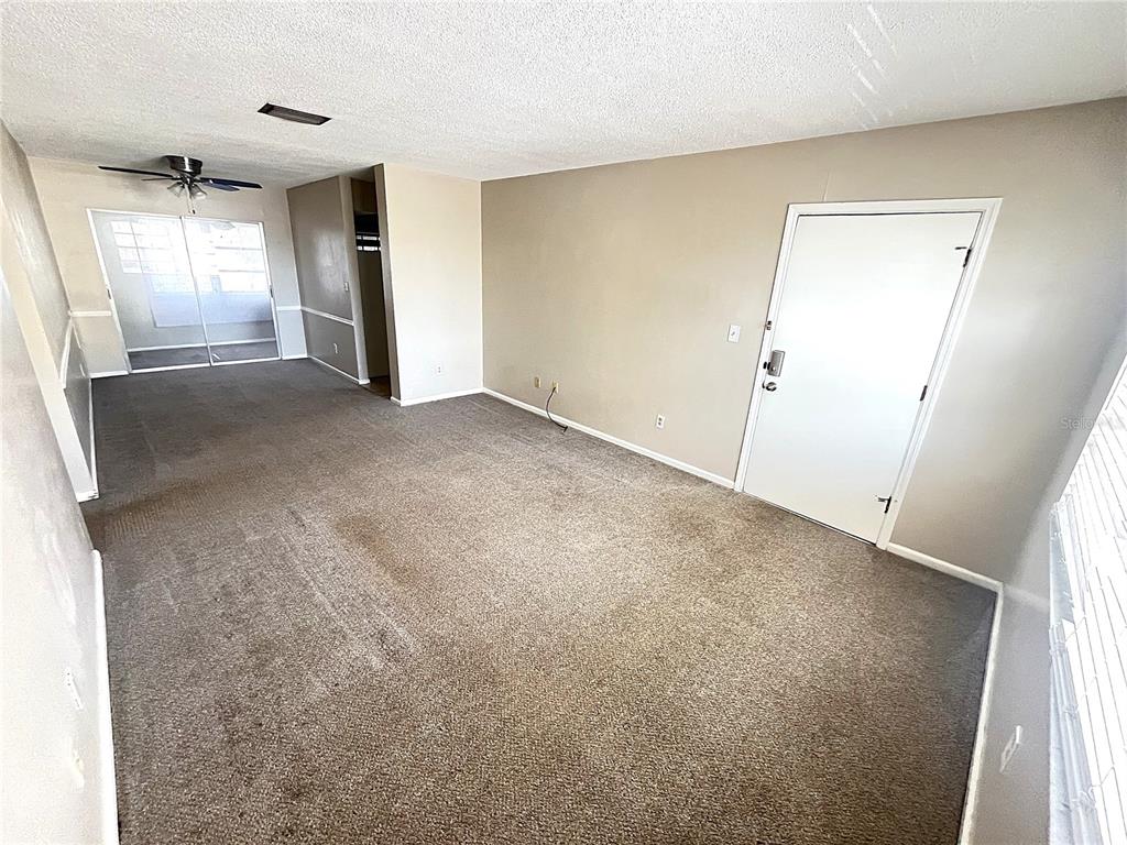 An empty living room features beige walls, brown carpeting, a ceiling fan, and a white door — 4507 9TH STREET W UNIT J6, BRADENTON, FL