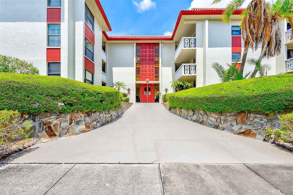 A concrete driveway leads to the red-trimmed entrance of a multi-story apartment building — 2612 PEARCE DRIVE UNIT 209, CLEARWATER, FL
