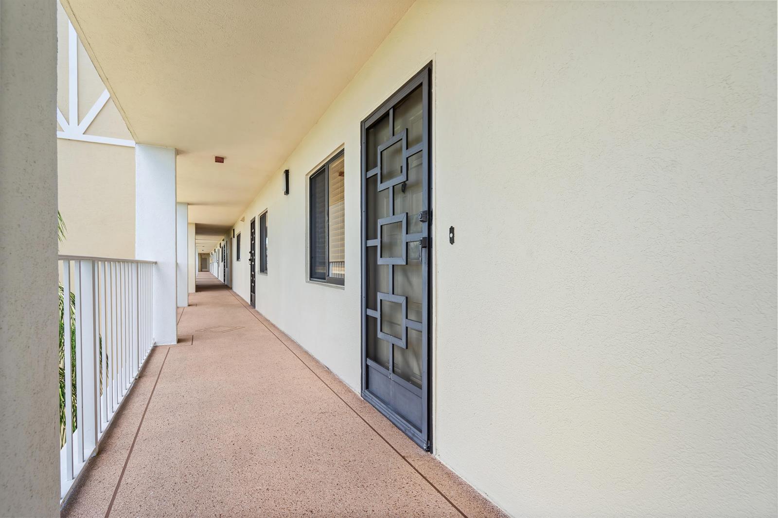 An outdoor apartment building walkway with a beige floor, white walls, and metal screen doors — 7290 Kinghurst Drive # 607, Delray Beach FL 33446, DELRAY BEACH, FL