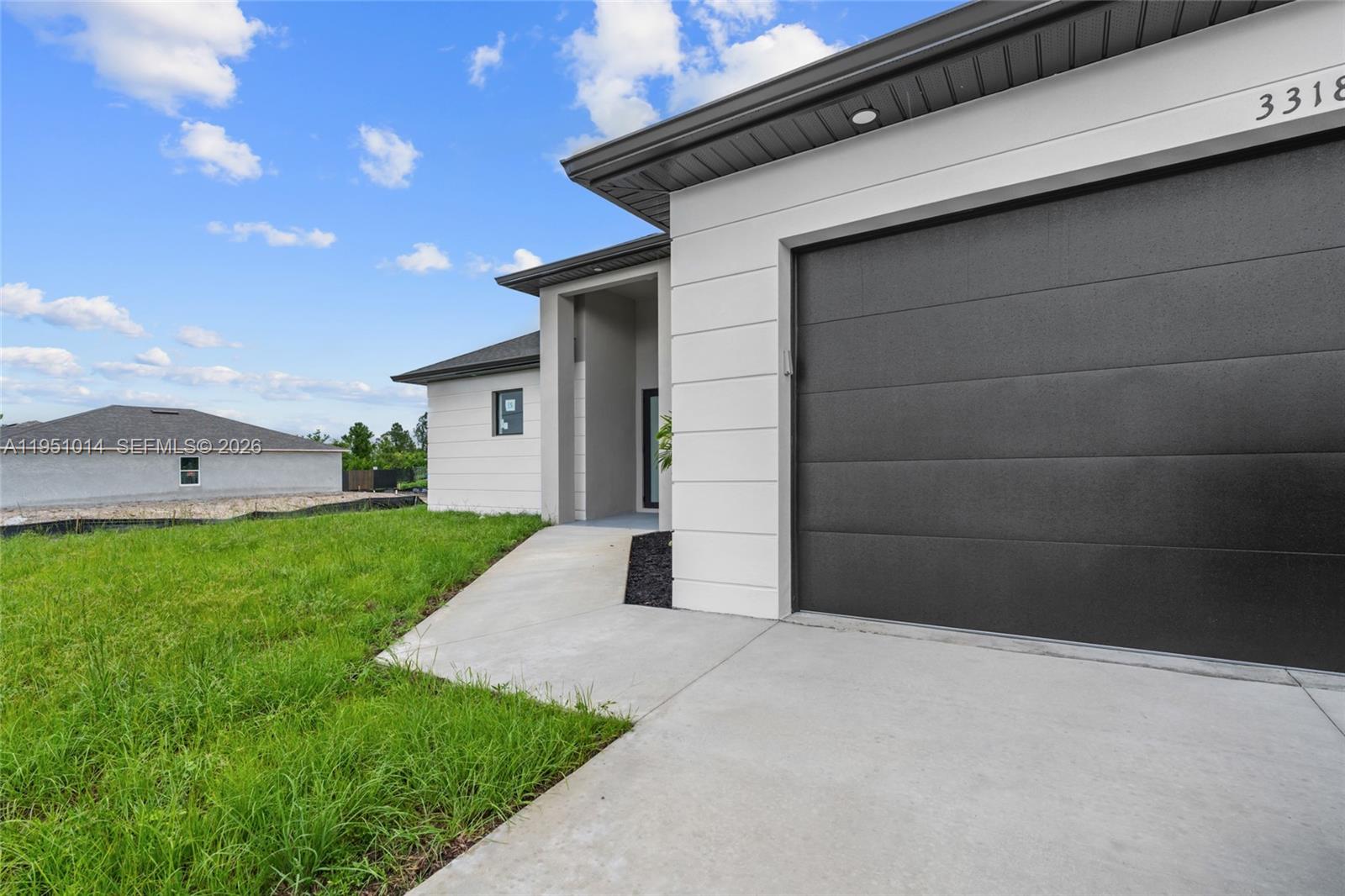 A modern house exterior features a dark garage door, white horizontal siding, and concrete walkway — 3318 30th St SW, Lehigh Acres FL 33976, LEHIGH ACRES, FL