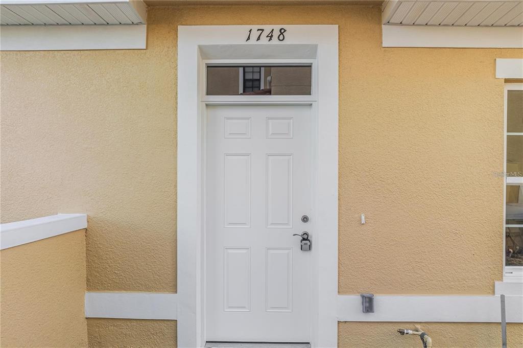 A white front door with a transom window is centered on a tan stucco exterior wall — 1793 CORIANDER DRIVE, POINCIANA, FL