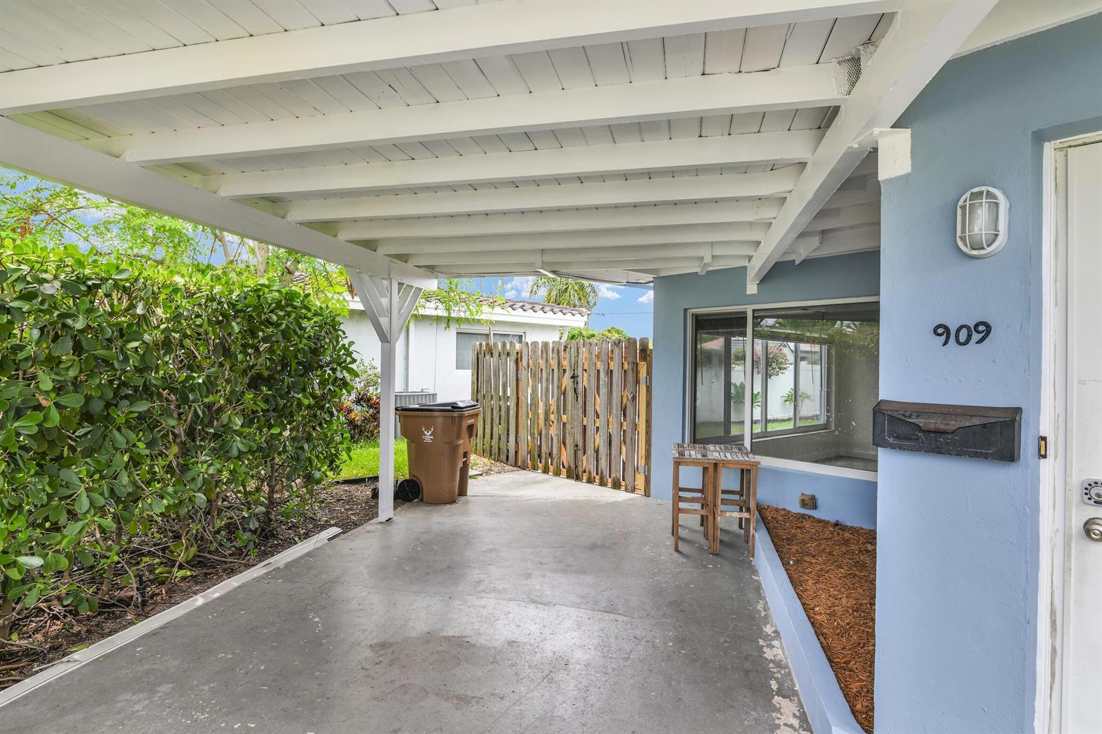 A covered concrete carport sits beside a light blue house exterior with a wooden fence — 909 SE 16 Place, Deerfield Beach FL 33441, DEERFIELD BEACH, FL