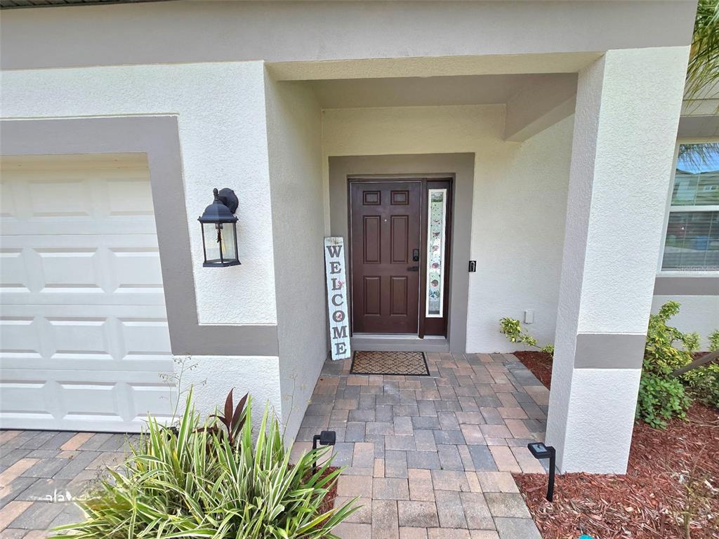 A brown front door with a side window sits under a covered entryway with pavers — 2950 ADAMS CROSS DRIVE, ZEPHYRHILLS, FL