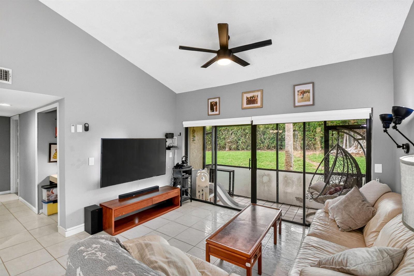 A living room features a vaulted ceiling, tile flooring, a wall-mounted television, and sliding doors — 14 Via De Casas Sur # 104, Boynton Beach FL 33426, BOYNTON BEACH, FL