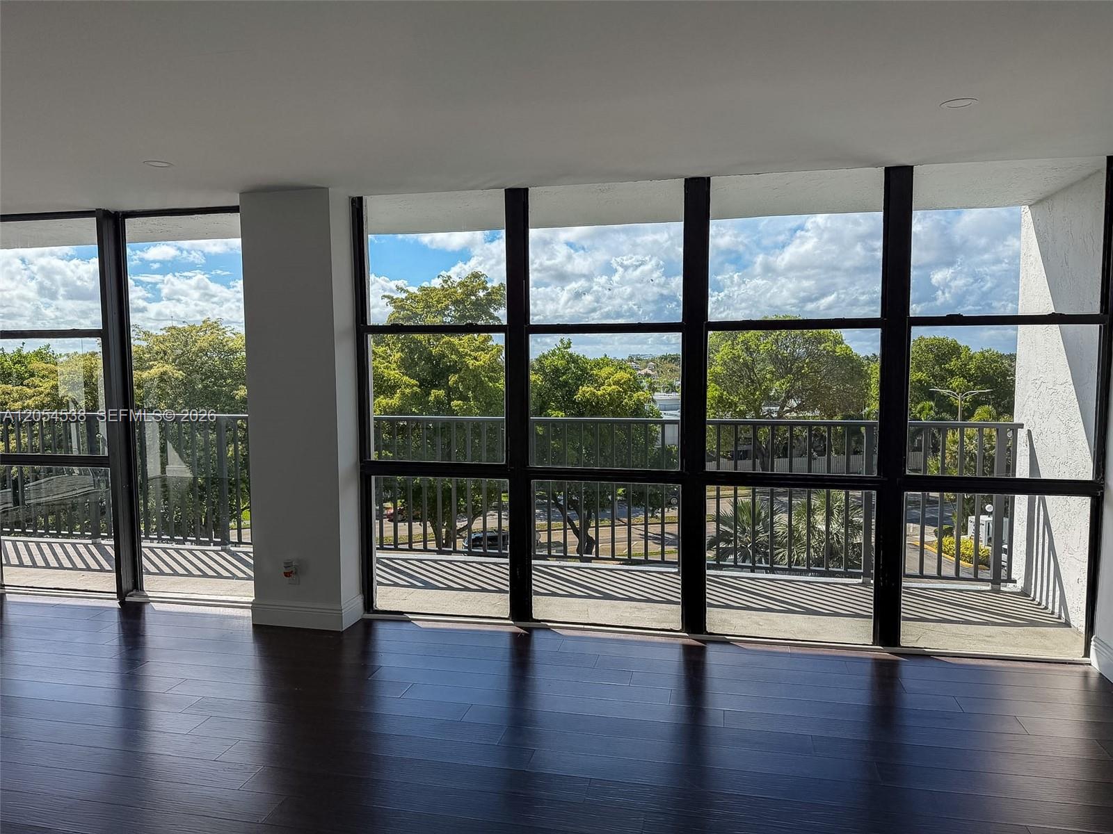 An empty room with dark wood flooring and large windows overlooking trees and a balcony — 1000 Parkview Dr # 531, Hallandale Beach FL 33009, HALLANDALE BEACH, FL