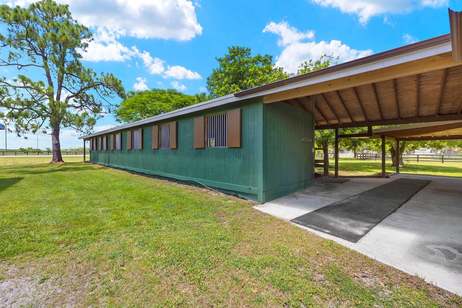 A green horse barn with a covered walkway and barred windows sits on a grassy lot — 16031 Rustic Road, Wellington FL 33470, WELLINGTON, FL