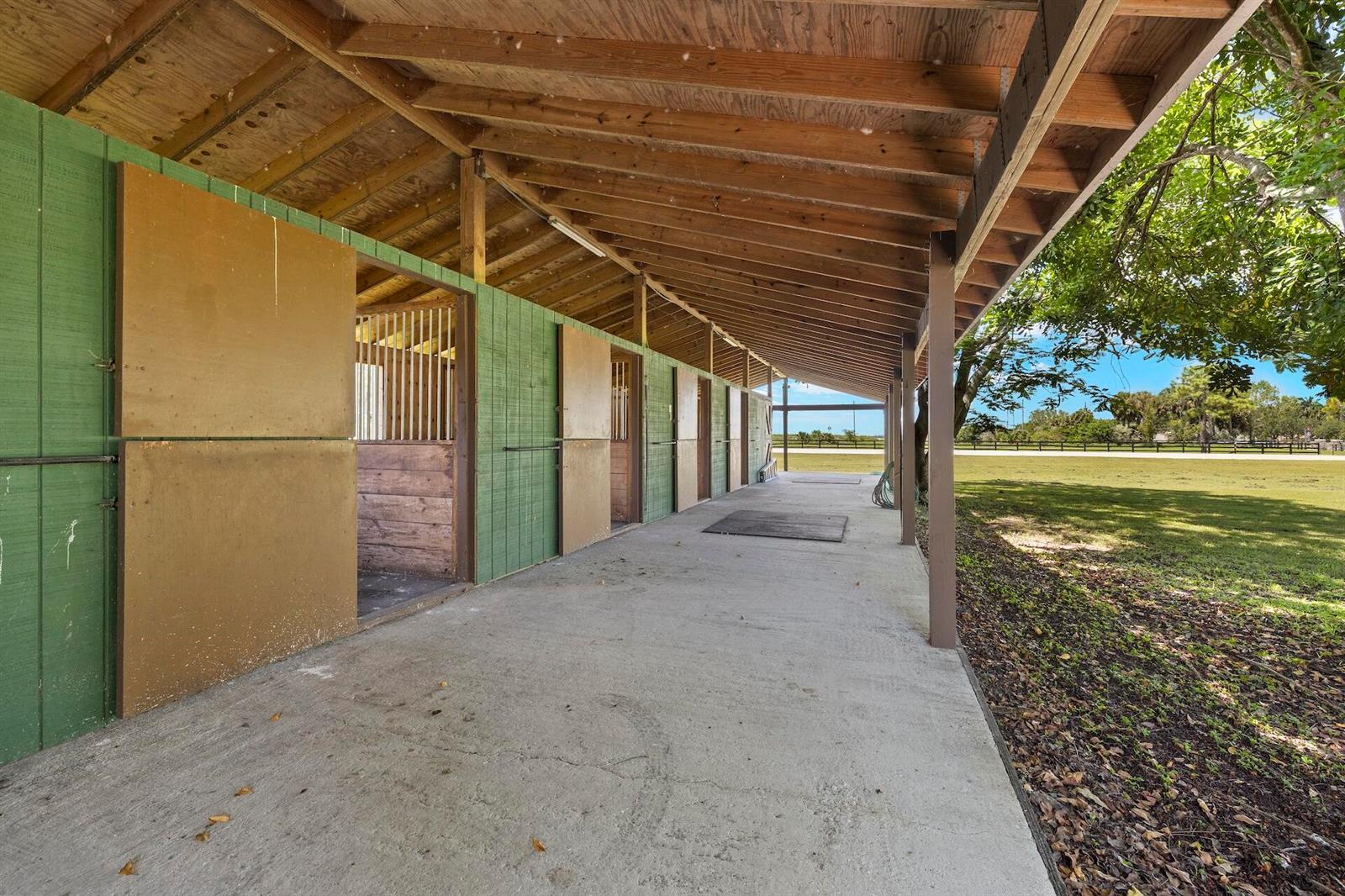 A covered concrete walkway runs alongside a row of horse stalls with green wooden doors — 16031 Rustic Road, Wellington FL 33470, WELLINGTON, FL