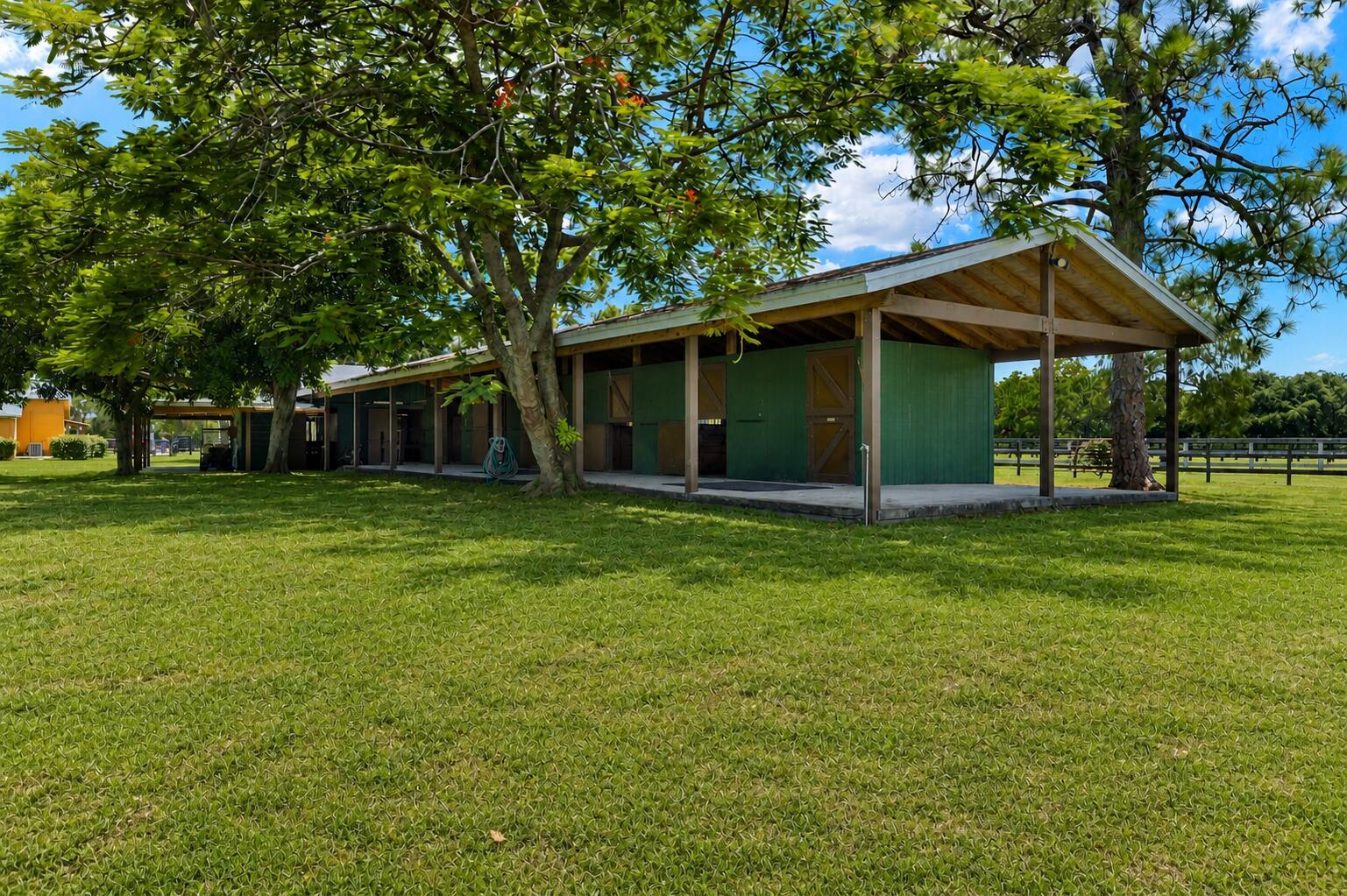 A long, green-sided horse stable with a covered porch sits on a grassy lawn — 16031 Rustic Road, Wellington FL 33470, WELLINGTON, FL