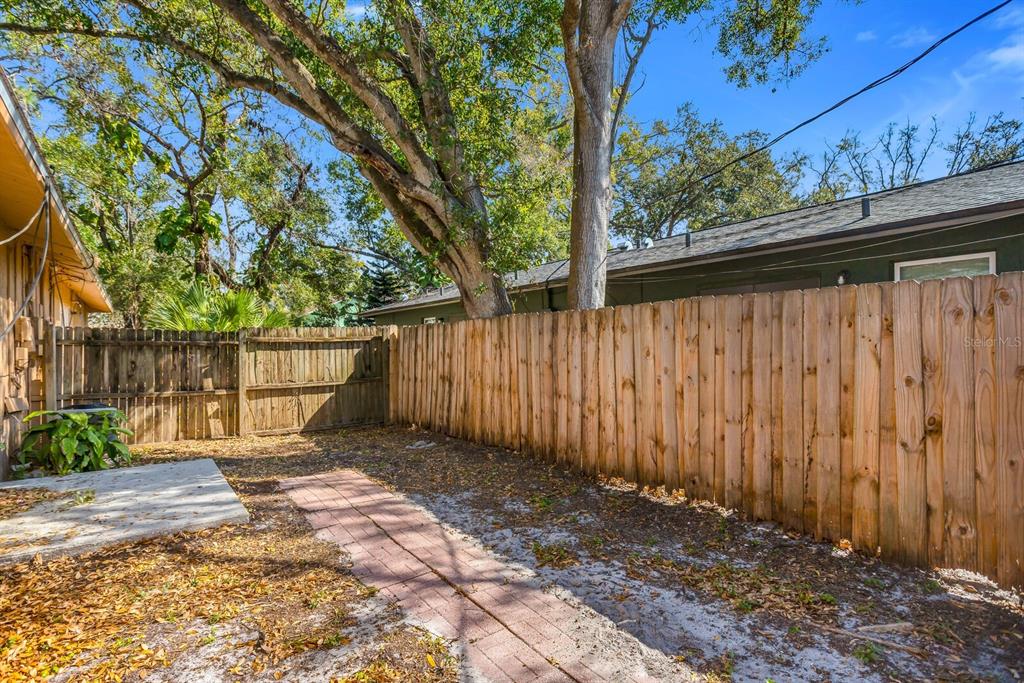A fenced backyard features a brick walkway, a concrete patio, and mature trees — 14717 SUNSET ST, CLEARWATER, FL