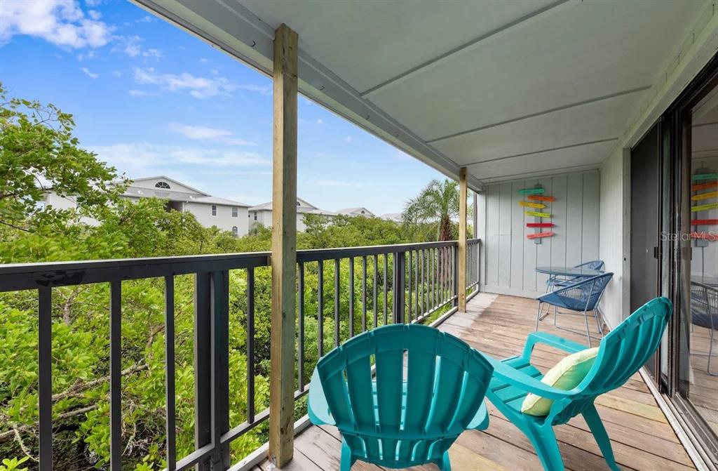A covered balcony features wooden flooring, black railings, and several chairs overlooking a wooded area — 19823 GULF BOULEVARD UNIT 42, INDIAN SHORES, FL