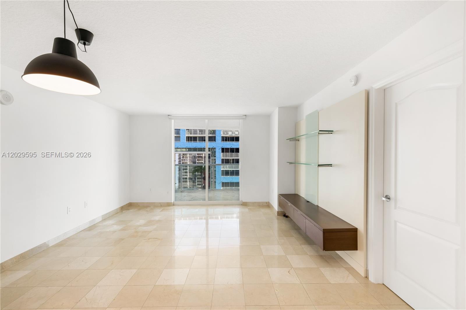 An empty room features beige tile flooring, a wall-mounted media console, and a balcony view — 2101 Brickell Ave # 1103, Miami FL 33129, MIAMI, FL