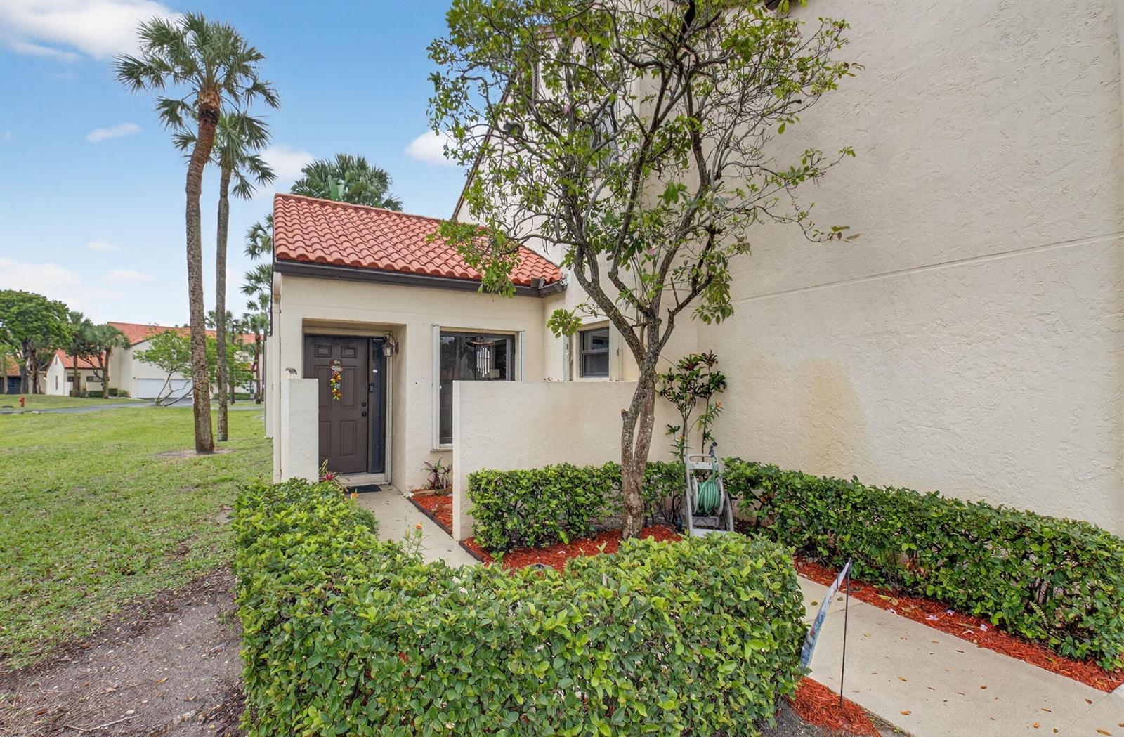 An exterior view of a townhouse entrance with a brown door, tiled roof, and landscaping — 10 Vía De Casas Sur # 101, Boynton Beach FL 33426, BOYNTON BEACH, FL