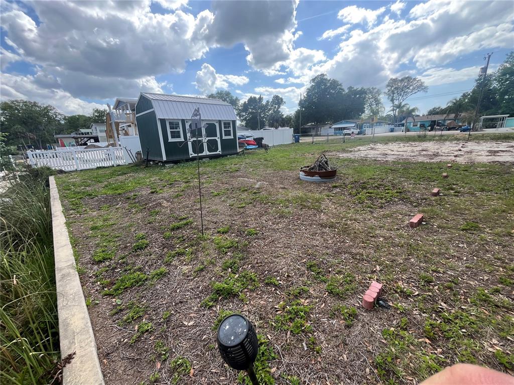 A backyard features a small shed, a fire pit, and a white fence under a cloudy sky — 7206 BETHESDA COURT, WEEKI WACHEE, FL