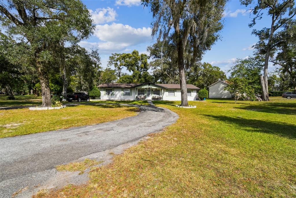 A single-story house with a curved driveway sits on a large, tree-filled grassy lot — 25309 CARNOUSTIE DRIVE, SORRENTO, FL