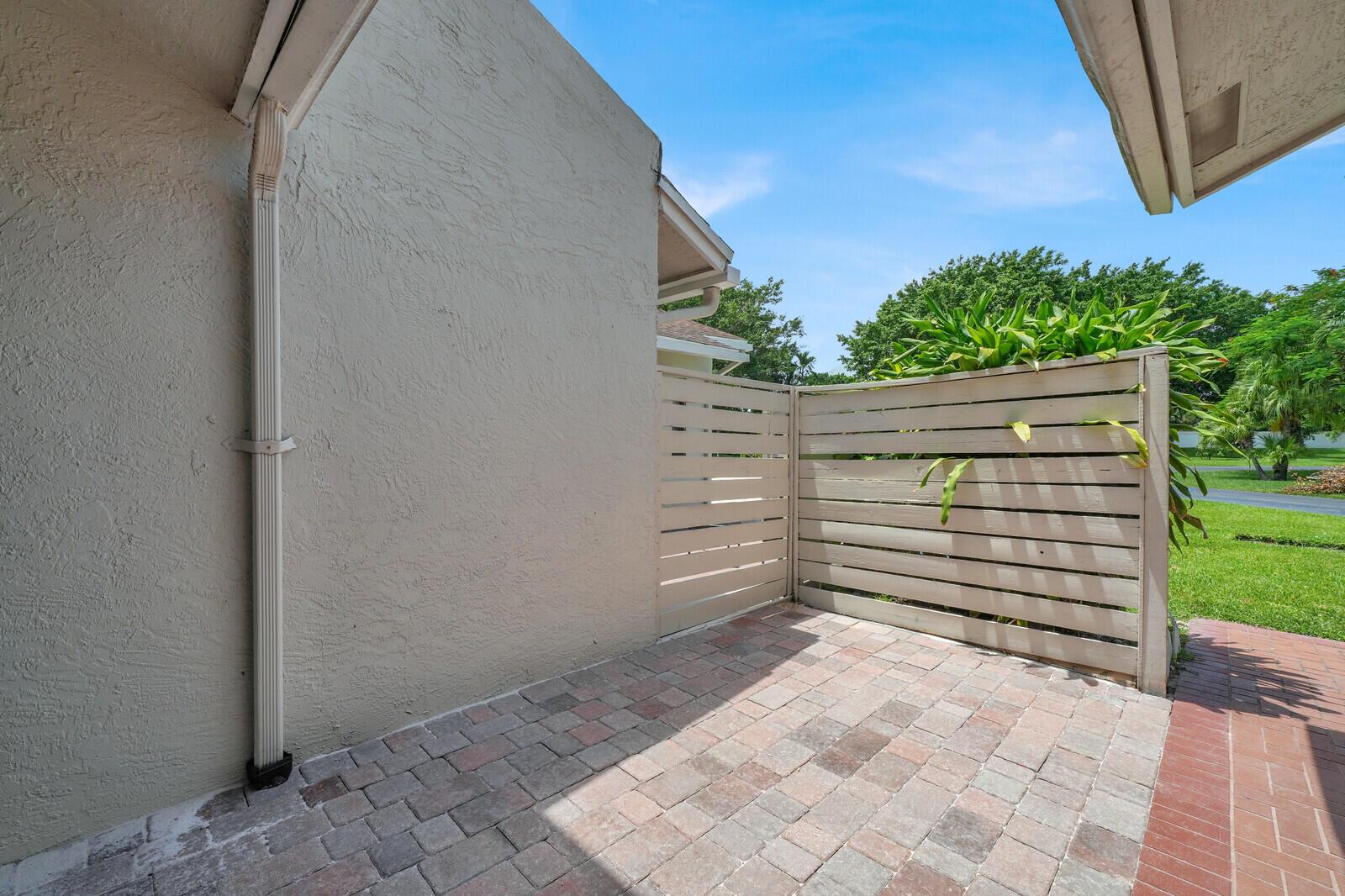 A paved patio area features a beige stucco wall, a downspout, and a horizontal privacy fence — 6371 Toulon Drive, Boca Raton FL 33433, BOCA RATON, FL