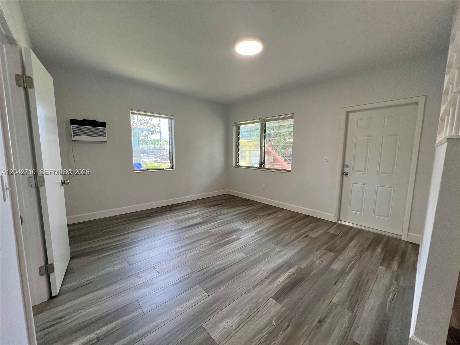 An empty room features gray wood-look flooring, white walls, two windows, and a white door — 962 NW 6th St # 1, Miami FL 33136, MIAMI, FL
