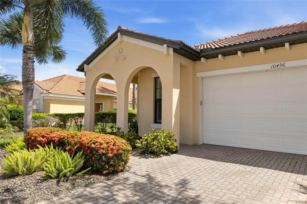 A beige house exterior features a two-car garage, arched entryway, and a brick paver driveway — 10496 CROOKED CREEK DRIVE, VENICE, FL