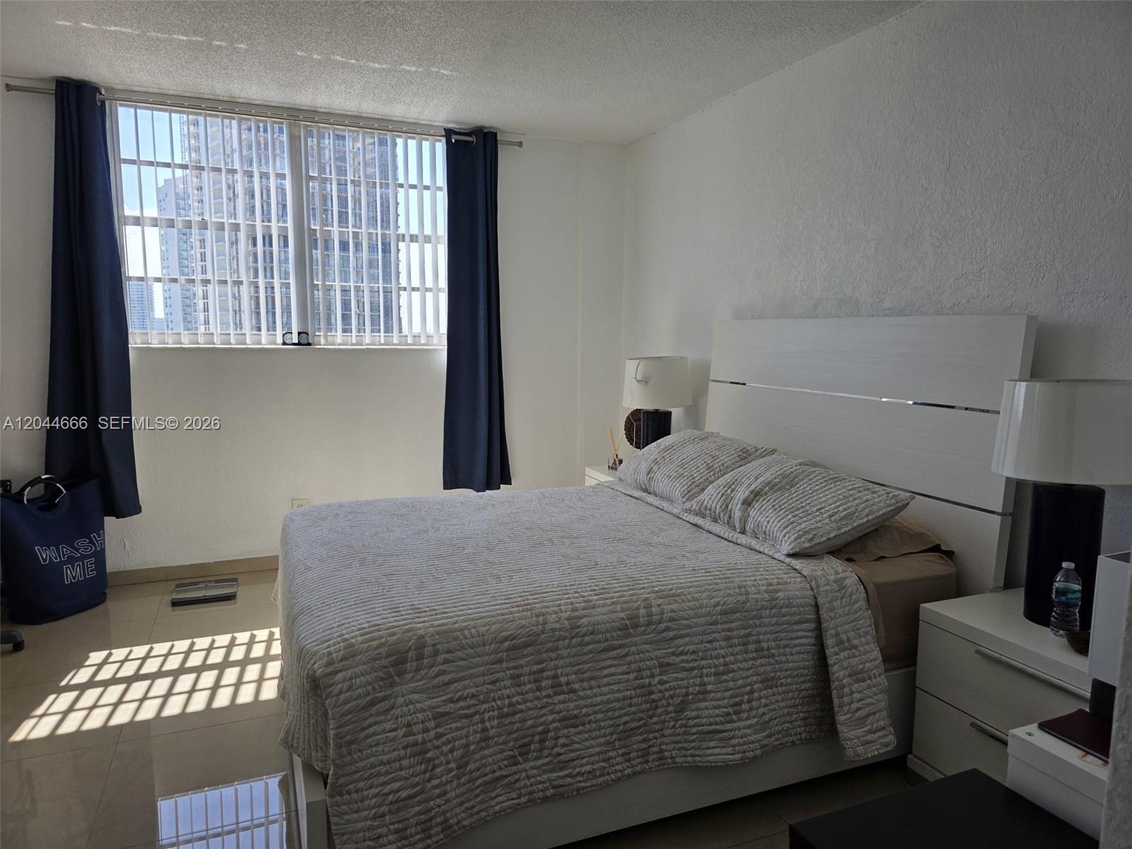 A bedroom features a white bed frame, two nightstands, and a window with blue curtains — SUNNY ISLES BEACH, FL