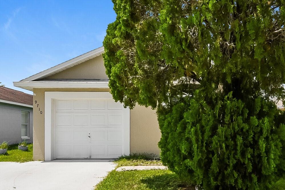 A beige house exterior features a white garage door and a large evergreen tree — 9710 RED CLOVER AVENUE, ORLANDO, FL