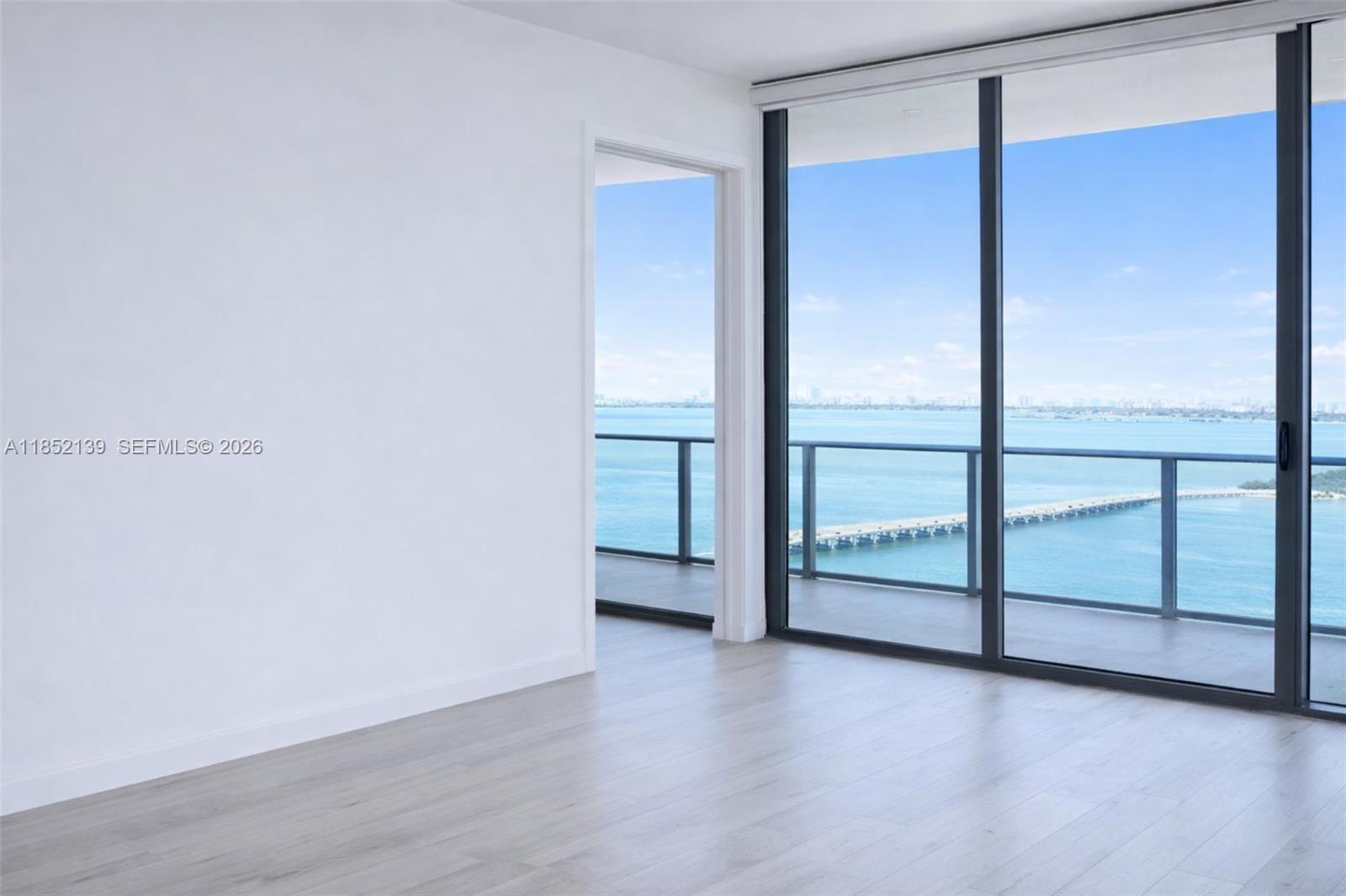 An empty room with light wood floors features floor-to-ceiling windows overlooking a bay and bridge — 480 NE 31st St # 3301, Miami FL 33137, MIAMI, FL