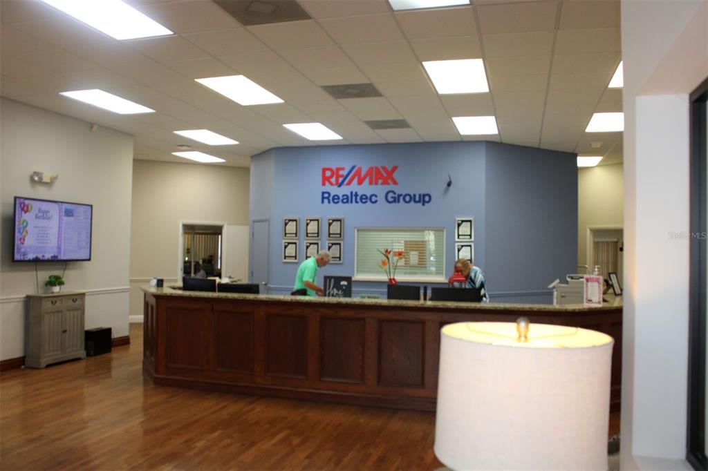 A reception area features a wooden desk, a blue wall with a Re/Max logo, and wood flooring — 4175 WOODLANDS PARKWAY UNIT 26, PALM HARBOR, FL