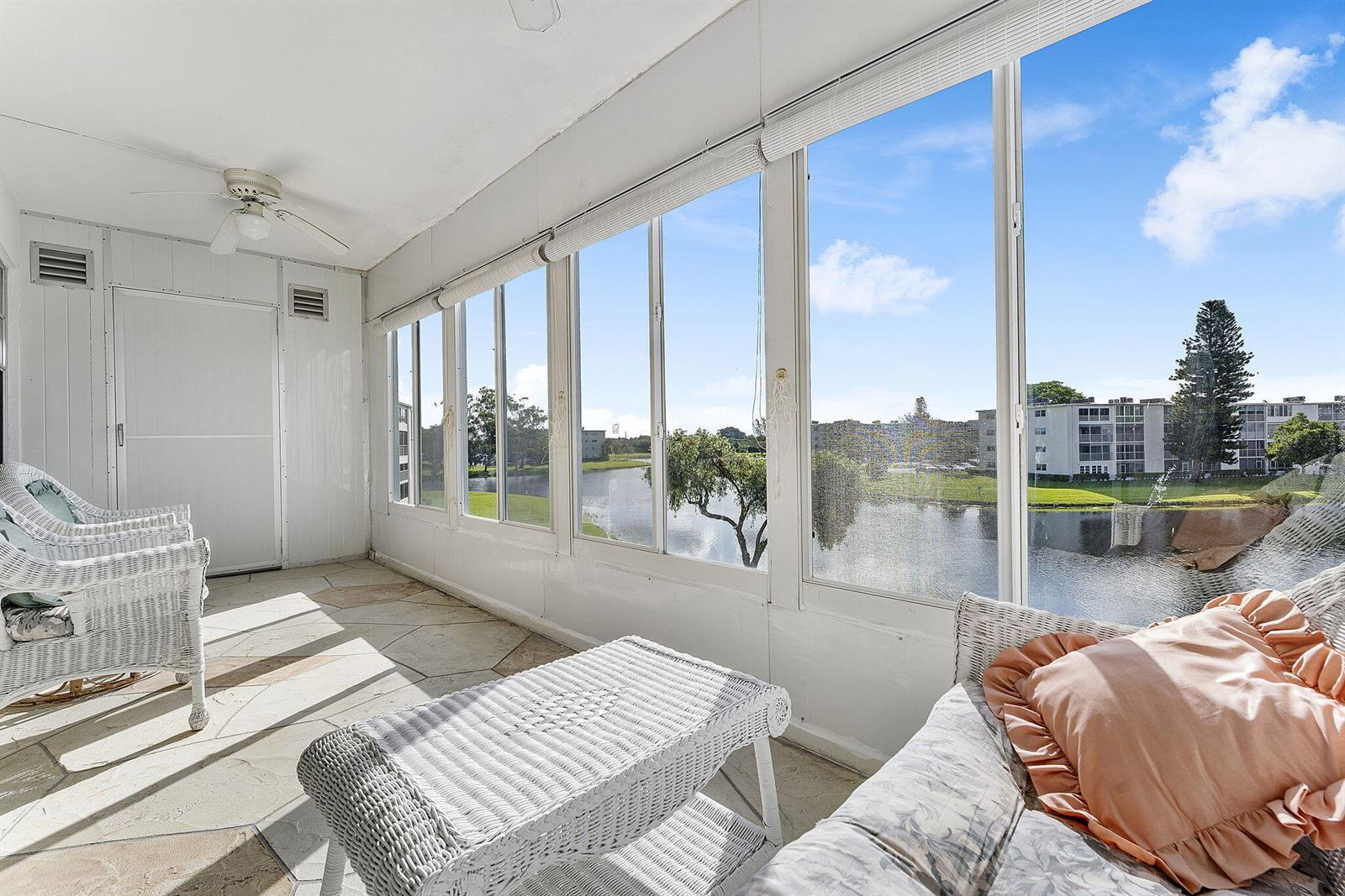 A sunroom with white wicker furniture, stone flooring, and large windows overlooking a lake — 3027 Guildford B, Boca Raton FL 33434, BOCA RATON, FL