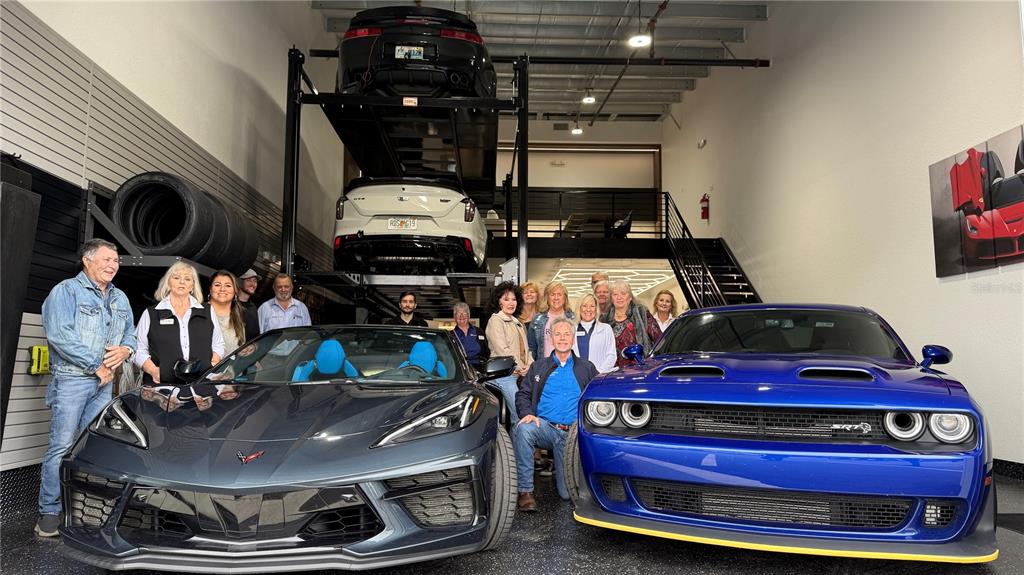 A group of people stands in a large garage featuring a car lift and sports cars — 8185 APEX WAY, SEBRING, FL