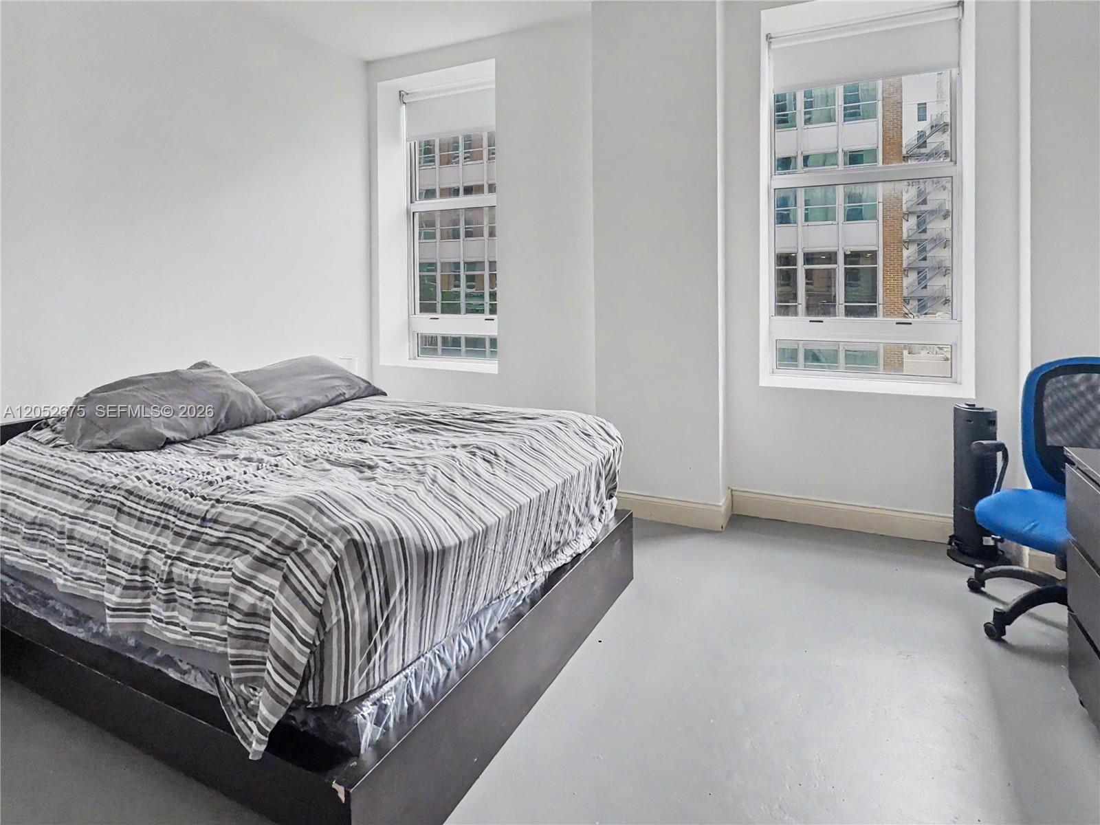A bedroom features a bed with striped bedding, two windows, and a blue office chair — 111 E Flagler St # 410, Miami FL 33131, MIAMI, FL
