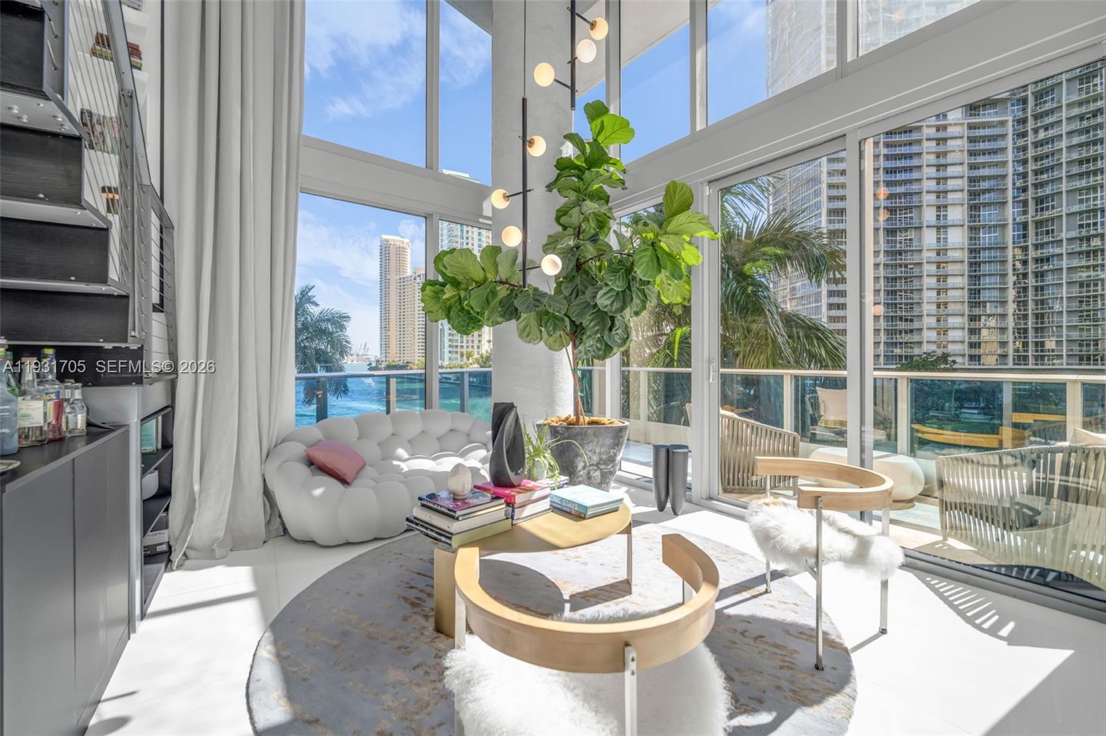 A modern living room features floor-to-ceiling windows, a large potted plant, and waterfront city views — 200 Biscayne Boulevard Way # 309, Miami FL 33131, MIAMI, FL
